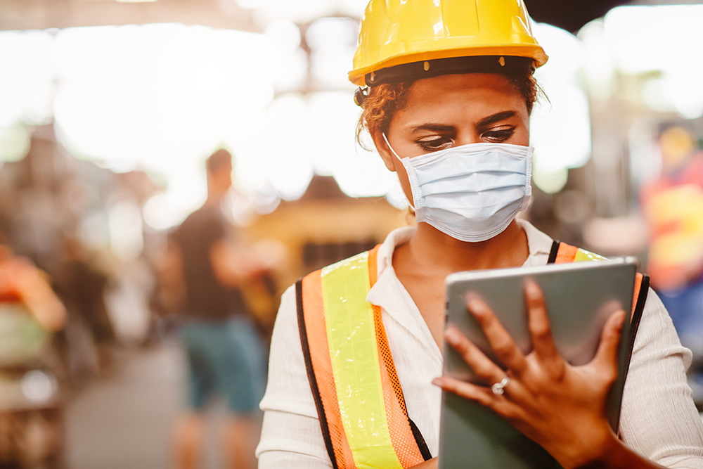 woman in high vest vest, hard hat and face mask looking at hand-held tablet