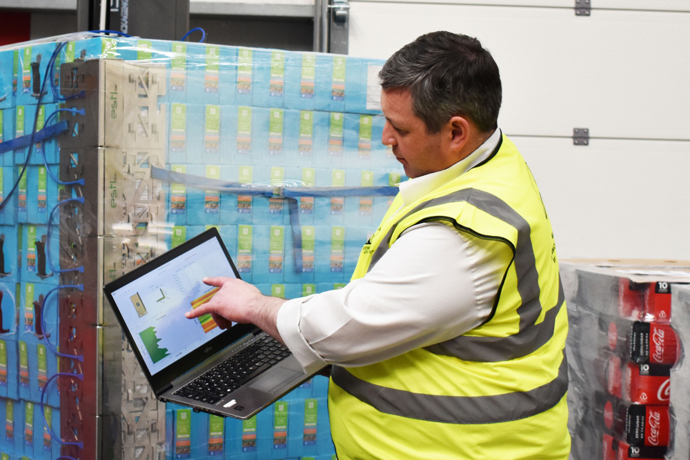 Man in hi-viz with laptop standing in front of pallet of wrapped boxes